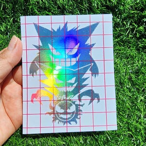 Könnte beinhalten: Ein holografischer Aufkleber mit einer farbenfrohen Illustration von Gengar und Gastly. Das Design befindet sich auf einem weißen Hintergrund mit einem roten Rastermuster. Der Aufkleber wird vor einem Hintergrund aus grünem Gras gehalten.