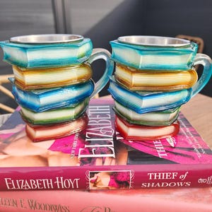 May include: Two decorative mugs shaped like stacks of books, with a silver rim and clear handles. The mugs are in shades of blue, yellow, and red. They sit atop a stack of books with the titles "Elizabeth Hoyt" and "Thief of Shadows".
