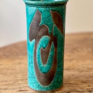 Könnte beinhalten: Eine türkisfarbene Keramikvase mit Craquelé-Glasur und braunem, abstraktem Design. Die zylindrische Vase hat einen leicht ausgestellten Rand und ist etwa 20 cm hoch.