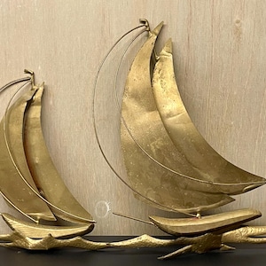 以下が含まれることがあります： 2つの金色の金属製ヨットの壁掛けアート。各ヨットは湾曲した帆とボートのベースを備え、細い金属線が上方に伸びています。ヨットは木製の背景に取り付けられています。
