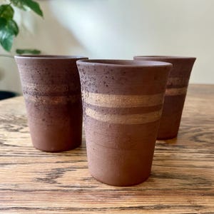May include: Three brown ceramic tumblers with horizontal gold bands. The tumblers have a textured surface and a slightly flared rim. They are arranged on a wooden surface, with a plant visible in the background.