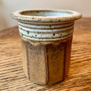 May include: A small, brown and white ceramic vase with a textured surface. The vase has a wide rim and a rounded base.