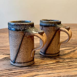 May include: Two handmade ceramic mugs with brown and blue glaze. The mugs have unique, asymmetrical handles.