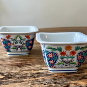 Könnte beinhalten: Zwei quadratische, weiße Keramikschalen mit farbenfrohen Blumen- und geometrischen Mustern. Die Schalen haben eine leicht ausgestellte Form und sind mit roten, orangefarbenen, grünen und blauen Designs verziert. Die Schalen stehen auf einer Holzoberfläche.