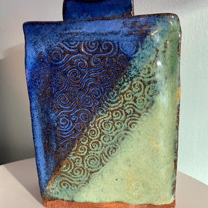 Puede incluir: Un jarrón de cerámica con un esmalte azul, verde y marrón. El jarrón tiene un diseño único de espiral y una forma rectangular.