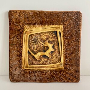 Könnte beinhalten: Quadratische Keramik-Wandkunst mit einem strukturierten braunen Rand und einem zentralen Quadrat mit einem wirbelnden, abstrakten Design in Braun- und Goldtönen. Das Kunstwerk hat ein rustikales, handgefertigtes Aussehen.
