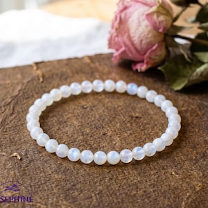 May include: A delicate white moonstone bead bracelet. The bracelet is strung on an elastic band and rests on a vintage, brown, textured surface. Dried pink roses are in the background. The bracelet is a beautiful piece of jewelry.
