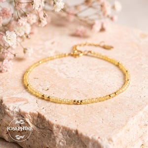 May include: A delicate bracelet featuring small, faceted yellow beads interspersed with tiny gold-toned metal spheres. The jewelry piece includes a gold-toned chain extension with a clasp and a small circular logo tag reading JOSEPHINE.