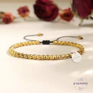 Könnte beinhalten: Ein goldenes Perlenarmband mit einem kleinen silbernen Anhänger, auf dem "Josephine" steht. Das Armband ist mit einer schwarzen Schnur gebunden und hat einen verschiebbaren Knoten zum Anpassen der Größe.