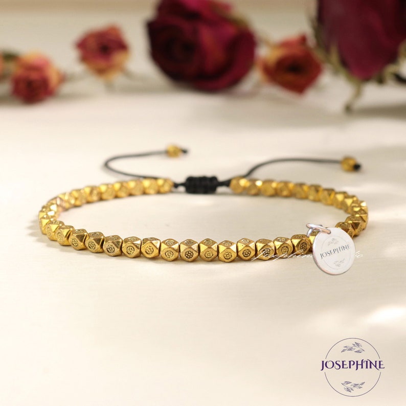 May include: A gold beaded bracelet with a small silver charm that says "Josephine" on it. The bracelet is tied with a black cord and has a sliding knot for adjusting the size.