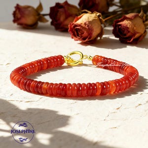 May include: A red beaded bracelet with a gold clasp. The bracelet is made of small, round, red beads. The clasp is a gold-colored ring. The bracelet is on a white surface. The brand name "Josephine" is visible.