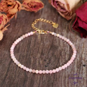 Könnte beinhalten: Ein zartes Armband aus rosa Opalperlen mit einem goldenen Herz-Anhänger und einem goldenen Kettenverschluss. Das Armband liegt auf einer Holzoberfläche.