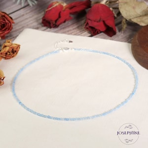 May include: A delicate, light blue beaded necklace with a silver clasp. The necklace is displayed on a white surface, with the brand name "JOSEPHINE" in a circular logo. Red roses and dried flowers are in the background.