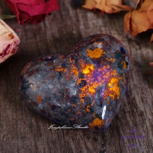以下が含まれることがあります： 灰色、オレンジ、紫の独特な大理石模様のハート型の研磨石。石は光沢のある仕上げで、暖色系の光に照らされています。