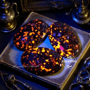 May include: Three polished, oval-shaped stones with a dark base and vibrant orange and yellow inclusions. The stones are arranged on a vintage book, with the text "Josephine Studio" visible. The overall aesthetic is mystical and eye-catching.