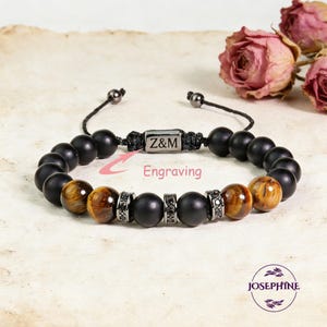 May include: A beaded bracelet featuring black matte onyx and tiger eye stones. The bracelet includes a metal accent piece with the initials "Z&M" and the word "Engraving". The bracelet is adjustable with a black cord.