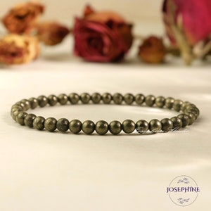 May include: A gold-toned pyrite beaded bracelet with small, round beads. The bracelet is lying on a white surface.