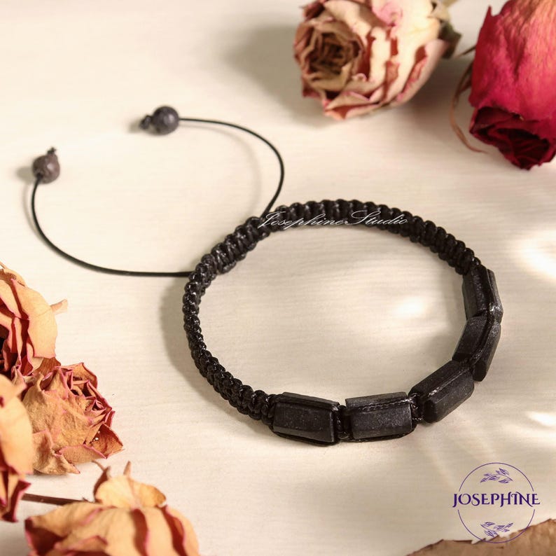 May include: Two black bracelets. One is a simple cord with a small bead at each end. The other is a braided bracelet with rectangular black beads. The image includes dried roses and the text "Josephine Studio".