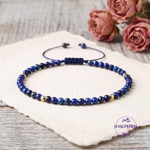 May include: A blue beaded bracelet featuring small faceted lapis lazuli stones and polished gold-tone metal beads. The bracelet has an adjustable cord closure with a sliding knot mechanism and two small beads at the ends of the pull strings.