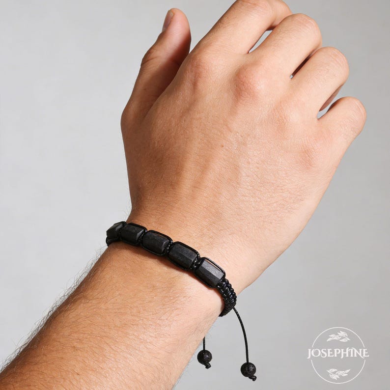 May include: Black beaded bracelet with rectangular beads and a woven cord. The bracelet is on a wrist. The bracelet has a sliding knot closure. The brand name "JOSEPHINE" is in the lower right corner.