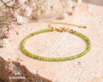 May include: A delicate bracelet featuring small, faceted green peridot beads strung on a thin gold-toned chain with a lobster clasp. The jewelry piece is displayed on a textured stone surface near small white and pink flowers.