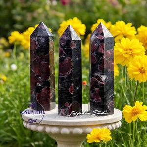 Op de afbeelding: Drie zwarte en rode granaat kristal obelisken met puntige toppen. De kristallen worden tentoongesteld op een witte sokkel in een tuin met gele bloemen. De kristallen hebben een gepolijste afwerking en een natuurlijk, gemarmerd uiterlijk.