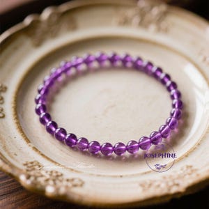May include: A purple amethyst beaded bracelet. The bracelet is made of small, round, polished amethyst beads. The bracelet is resting on a white plate with a decorative gold rim. The plate has a floral design.
