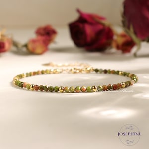 May include: A delicate gold chain bracelet with small green and red gemstone beads. The bracelet is on a white surface with dried roses in the background.