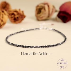 May include: A delicate hematite anklet featuring small, faceted black beads strung together. The anklet has a silver-colored clasp and is displayed on a light beige surface. The text "• Hematite Anklet •" is below the anklet.