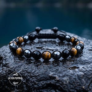May include: A beaded bracelet featuring alternating round tiger eye, polished black onyx, and metallic hematite beads strung on a black cord. The design includes a sliding knot closure and a circular logo with the text JOSEPHINE at the bottom.