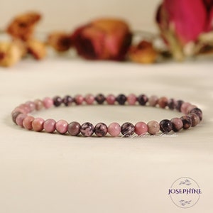 May include: A delicate bracelet composed of small, round, pink and dark brown beads. The beads are strung together to form a flexible band. The background features blurred dried flowers, adding a soft, natural aesthetic.