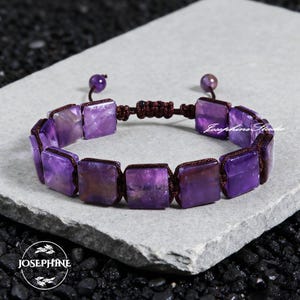 May include: A purple amethyst bracelet with square-cut stones. The stones are connected by a brown cord, and the bracelet has a sliding knot closure. The bracelet is displayed on a gray stone surface. The brand name "Josephine Studio" is visible.