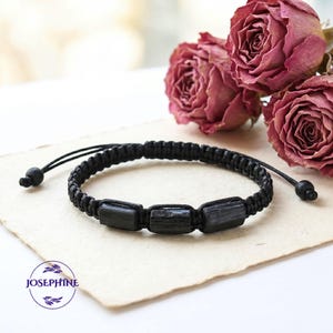 May include: Black braided bracelet featuring three rectangular black stones. The bracelet has adjustable black cords with small, round beads at the ends. The bracelet is displayed on a cream-colored surface with dried pink roses in the background.