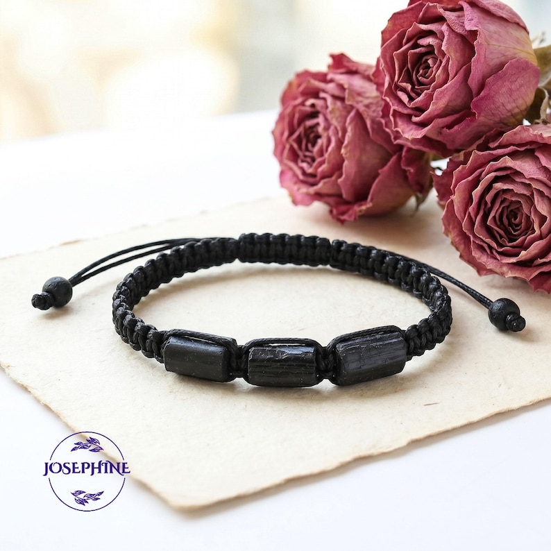 May include: Black braided bracelet featuring three rectangular black stones. The bracelet has adjustable black cords with small, round beads at the ends. The bracelet is displayed on a cream-colored surface with dried pink roses in the background.