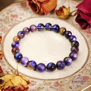 May include: A purple and gold tiger's eye beaded bracelet on a white plate with dried roses. The bracelet is made of round beads with a shimmering effect.
