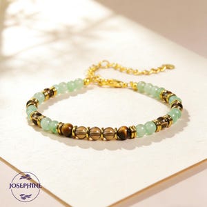 Puede incluir: Una pulsera de cuentas verdes claras y marrones con detalles dorados. La pulsera tiene una cadena y un cierre dorados. La pulsera está sobre una superficie blanca. La marca "JOSEPHINE" está en la esquina inferior izquierda.