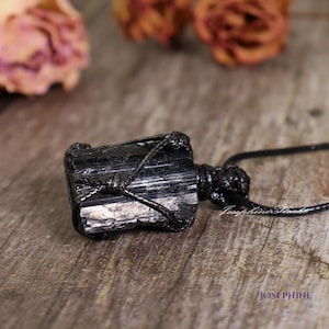 May include: A black tourmaline crystal pendant, wrapped in black cord, hangs on a black necklace. The rough-hewn stone has visible striations and is set against a blurred background with dried flowers. The word "JOSEPHINE" is visible in the lower right corner.