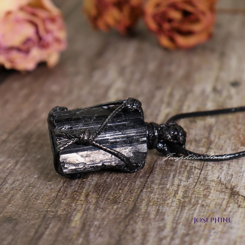 May include: A black tourmaline crystal pendant, wrapped in black cord, hangs on a black necklace. The rough-hewn stone has visible striations and is set against a blurred background with dried flowers. The word "JOSEPHINE" is visible in the lower right corner.