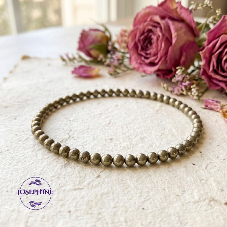 K&ouml;nnte beinhalten: Ein Armband aus silberfarbenen Perlen, bestehend aus kleinen, runden Perlen. Das Armband liegt auf einer strukturierten, cremefarbenen Oberfl&auml;che. Getrocknete rosa Rosen und andere Blumen befinden sich im Hintergrund. Der Markenname "JOSEPHINE" befindet sich unten links.