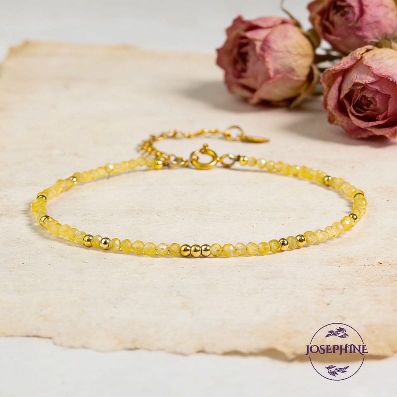 May include: A delicate bracelet featuring small, faceted yellow beads interspersed with polished gold-tone metal spheres. The piece includes a gold-tone metal chain extension with a circular clasp and a small tag. The text JOSEPHINE is visible.