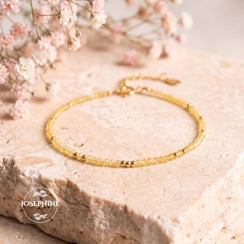 May include: A delicate bracelet featuring small, faceted yellow beads interspersed with tiny gold-toned metal spheres. The jewelry piece includes a gold-toned chain extension with a clasp and a small circular logo tag reading JOSEPHINE.