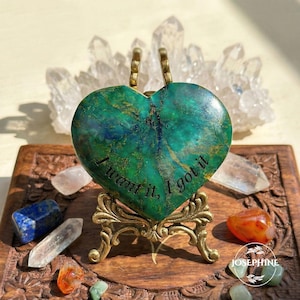 May include: A heart-shaped, green and gold stone with the text "I want it, I got it" on a decorative stand. The stone is displayed on a wooden carved box with crystals and gemstones.