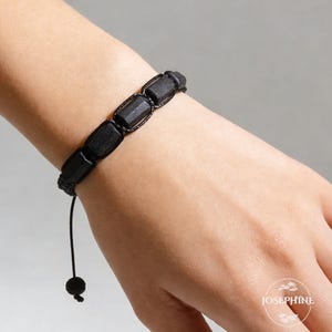 May include: Black beaded bracelet featuring rectangular beads. The bracelet is strung on a black cord with a sliding knot closure. The bracelet is shown on a wrist.