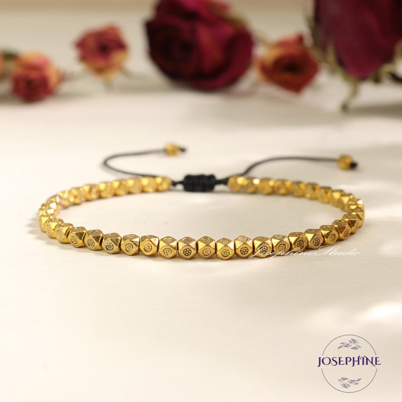 May include: A gold beaded bracelet with a black cord closure. The beads are small and have a geometric design.