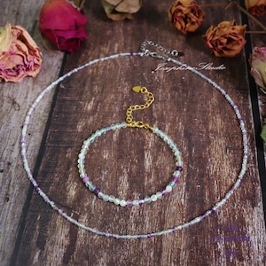 May include: A set of two necklaces made with small, colorful beads. The necklaces are made with light blue, purple, and white beads. The necklaces are on a wooden surface with dried roses.