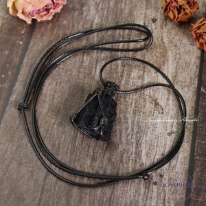 May include: A black tourmaline crystal pendant necklace. The raw, natural stone is encased in a woven wire setting and suspended from a black cord. The necklace is displayed on a wooden surface.