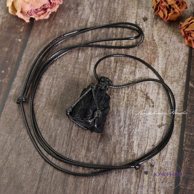 May include: A black tourmaline crystal pendant necklace. The raw, natural stone is encased in a woven wire setting and suspended from a black cord. The necklace is displayed on a wooden surface.