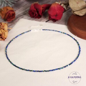 Peut inclure: Un collier délicat en chaîne argentée avec de petites perles de pierres précieuses bleues et vertes. Le collier est sur un fond de tissu blanc.