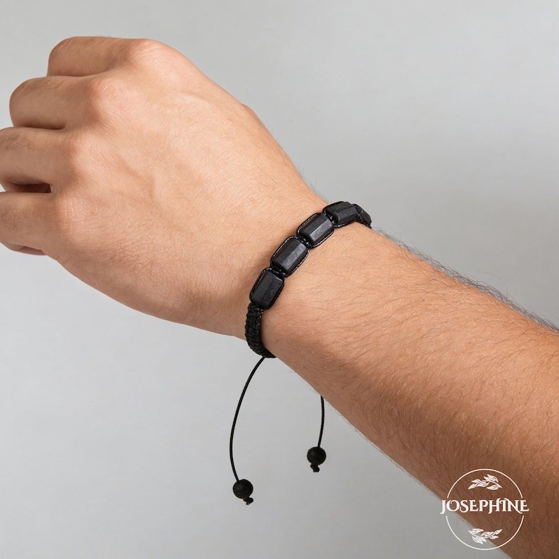 May include: Black beaded bracelet with rectangular beads. The bracelet is made of a woven black cord and has an adjustable closure. The bracelet is shown on a person's wrist.