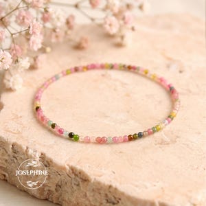 Può includere: Un braccialetto delicato composto da piccole perline rotonde di pietre preziose multicolori nelle tonalità del rosa, verde, giallo e blu. Il braccialetto è disposto in forma circolare e presenta il testo JOSEPHINE all'interno di un logo circolare.